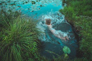 Büyük çimenler ve yapraklarla çevrili bir göle ya da göle atlayan bir kadın. Bir kadının arkası ve kıvırcık saçları suya girerken görülüyor..