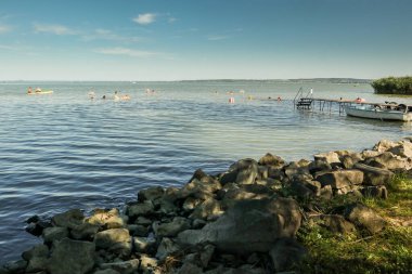 Macaristan 'da Balaton Gölü kıyısında kolay bir öğleden sonra. Sahilde ön planda yüzücüler ve arka planda su aktiviteleri olan kayalar. İskelede bir tekne görüldü. .