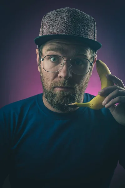 Hipster looking young male with glasses and hat is using a banana as a telephone, on a purple or pink background. Concept of a communication with banana.