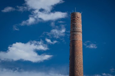 Güneşli bir günde, arka plan olarak mavi gökyüzü ve kabarık beyaz bulutlar olan uzun tuğlalı sanayi bacası.