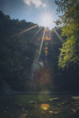 Slovenya 'da Divje Jezero adlı küçük bir gölün fotoğrafı. Gölün üzerinde güçlü bir güneş parlaması ve yüksek dikey kaya oluşumu var..