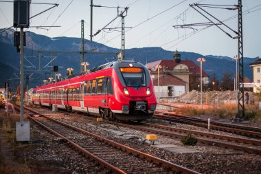 Kırmızı banliyö treni akşam saatlerinde Garmisch Partenkirchen istasyonuna giriyor. Dağların ve karlı tepelerin muhteşem arka plan manzarası var..