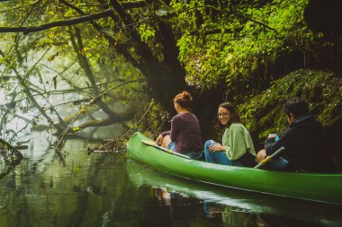 Üç kişilik bir grup, iki beyaz kadın ve bir erkek sisli ve soğuk suda sisli ve yeşil arka planda yeşil kanoyla kürek çekiyorlar..