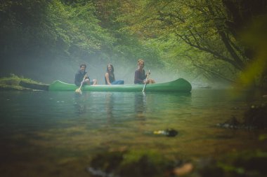 Üç kişilik bir grup, iki beyaz kadın ve bir erkek sisli ve soğuk suda sisli ve yeşil arka planda yeşil kanoyla kürek çekiyorlar..