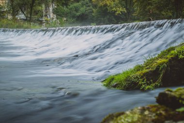 Slovenya 'nın Bela Krajina kentindeki Krupa nehri kaynağı veya baharının panoraması sisli bulutlu bir günde. Arka planda kaya oluşumuyla sisli yeşil bir nehir ve uzun mistik bir baraj ve sütlü suyun üzerinden akan hızlı su..