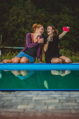 İki genç kadın havuzun yanında oturup içki içerken tezahürat yapıyor ve selfie çekiyor. Havuzda eğlenen ve gülümseyen kadınlar. Arkadaşlık ve mutluluk.