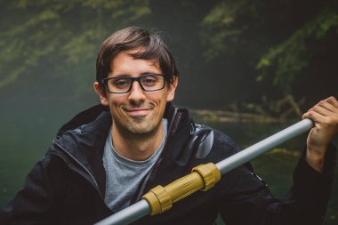 Siyah su geçirmez ceket giyen ve elinde kürek ve yeşil arka plan tutan siyah saçlı genç bir adamın portresi. Gülümseyen ama ağzı kapalı bir adam..