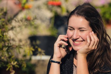 Gözleri kapalı rüya görürken telefonda gülümseyen ve konuşan beyaz bir kadın. Renkli bir bahçenin açık hava ortamı.