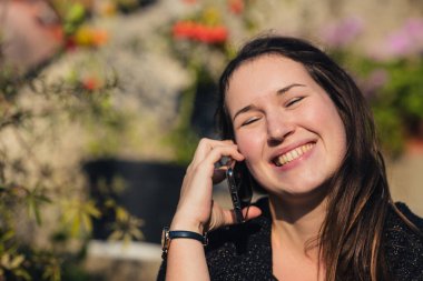 Gözleri kapalı kameraya bakarken telefonda gülümseyen ve konuşan beyaz bir kadın. Renkli bir bahçenin açık havası..