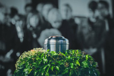 A metal urn with ashes of a dead person on a funeral, with people mourning in the background on a memorial service. Sad grieving moment at the end of a life. Last farewell to a person in an urn. clipart