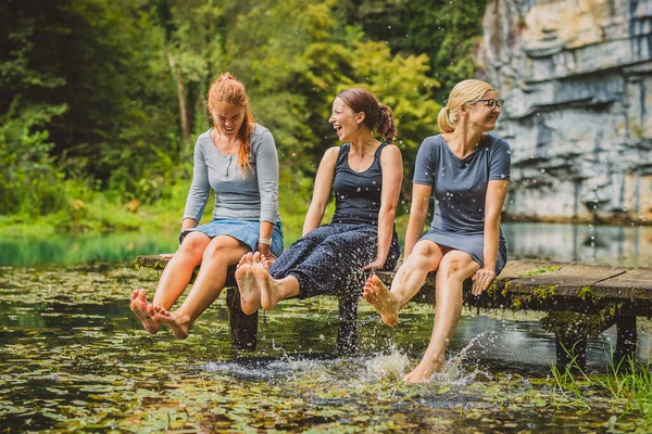 Three young women  relaxing on a wooden pier and splashing their feet into cold water. Picturesque clean lake with women splashing their feet in water. Concept of fun sexy outdoor activity.