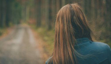 Mavi paltolu sarışın bir kız sık bir sonbahar ormanında orman yolunda yürüyor. Arkadan ya da arkadan ormandaki bir kadının görüntüsü.