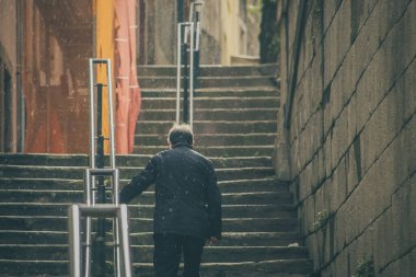 Bilinmeyen yaşlı bir adam yağmur sırasında merdivenlerden çıkıp parmaklık tutuyor. İnsan yağmur yağsa bile şemsiye takmaz..