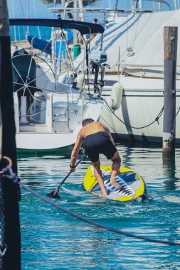 Park etmiş ya da demirlemiş tekneler arasında bir yat limanında SUP tahtasından denize düşen bir adamın arka görüntüsü. Sup tahtası sıralamasında oyunculuğu şaşırt