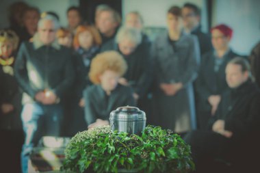 Cenaze töreninin arka planında yas tutan insanların külleri olan metal bir vazo. Bir hayatın sonunda kederli bir an. Vazodaki birine son bir veda..