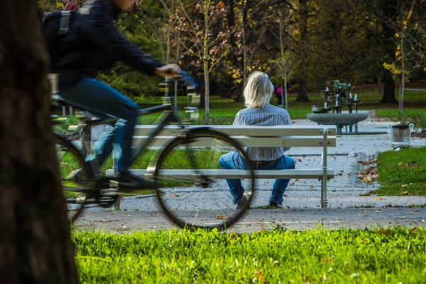 An older man with white hair and moustache enjoying a sit on the bench in a park with a fountain on an autumn sun while cyclist rush past him