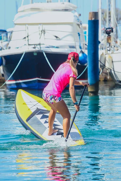 Sexy woman paddling on a SUP or stand up paddle board in a marina packed with boats. Girl turning a SUP board between yachts in a marina on a sunny day.