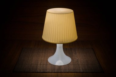 Ahşap arka planda, karanlık bir odada beyaz masa lambası. Sıradan, basit ama güzel plastik plastik plastik bir masa lambası tahta bir masanın üzerinde duruyor..