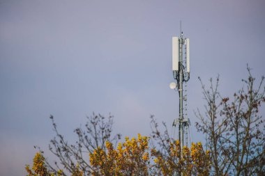 Kulede GSM antenleri, bulutlar, arka planda gökyüzü ve ön planda ağaçlar var. Anten için 5G 4G ve 3G LTE mobil kule. Haberleşme direği.