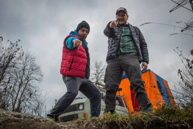 Kamp yaparken vahşi doğada poz veren spor elbiseli iki adam. Brokeback Mountain modern tarzı, kamerayı işaret eden eğlenceli bir çift. Campervan arkada..