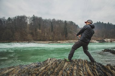 Nehrin kenarında açık havada beyzbol şapkalı genç bir adam balık tutuyor. Arka planda orman ve akan vahşi nehir.