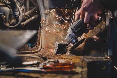 Bir arabanın gövdesindeki paslı metali kesip, onu restore etmek ve yeni bir parça haline getirmek için açı öğütücü kullanan bir adamın çıplak elleri. Kıvılcımlar uçuşuyor, aletler ve öğütücü tabaklar etrafta.