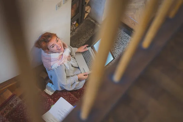 Young woman learning on the stairs next to a fireplace, holding a laptop and looking up. Romantic setting for learning in winter. Enjoying cozy winter learning session.