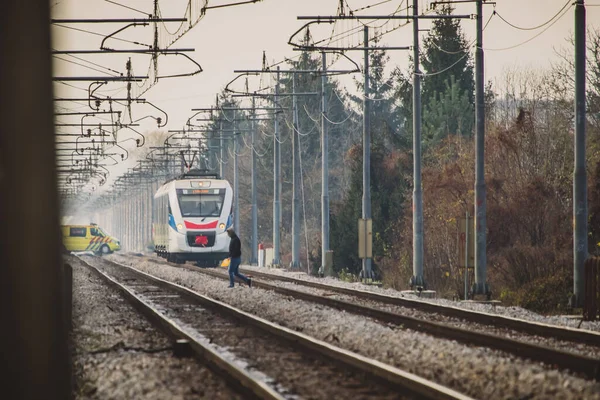 男は電車の線路を無視して白い旅客列車に近づいて歩く 救急車が列車の後ろの線路を渡っている 危険な軌道交差の概念 — ストック写真