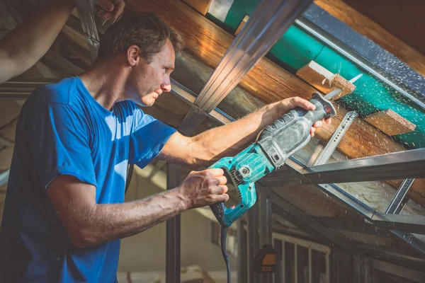 A person is cutting a board of wood next to a window with a reciprocating saw. Sawdust is flying around. Detail of a man cutting wooden board with strong back light to emphasize saw dust.