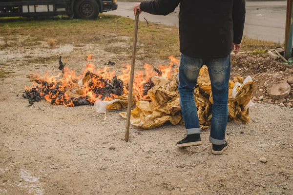 A man is burning a fire out of paper in an open space. Burning paper waste in residental or industrial environment. Pollution with burning paper.