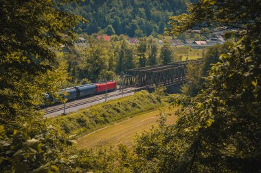 Kırmızı lokomotifli Avrupa yük treni, metal bir köprüye doğru giden iki raylı bir demiryolu hattında ilerliyor. Yapraklardaki bir delikten romantik bir manzara.