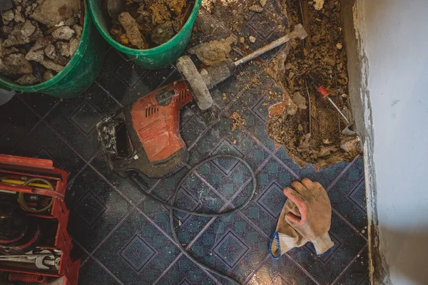 Flatlay photo of demolition hammer on the floor, open hole in the tiled floor, tools, old material and gloves next to an open hole with pipe are seen.