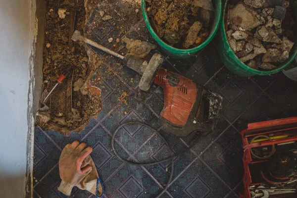 Red electric demolition hammer on the floor, while renovating an old water pipe in the ground. Construction site with tools, old material and gloves is seen.