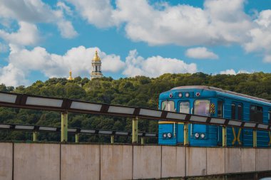 Kiev 'deki eski metro treni, Ukrayna' da sıcak bir yaz gününde Dinyeper nehri üzerinden Hydropark ve Dnipro bölgesine doğru ilerliyor. Arka planda Laura kiliseleri kompleksi görülüyor, Kiev simgesi..