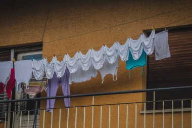 Eski beyaz iç çamaşırı ve diğer kıyafetler balkonda kuruyor, çamaşır ipine asılıyor, sarı bir cephenin önünde..