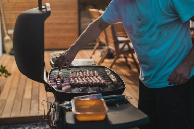 Balkan spesiyal yemeği ile ızgara tabağı hazırlayan bir adam var. Ahşap bir veranda ve çimenlik alanda açık sigara ızgarası.