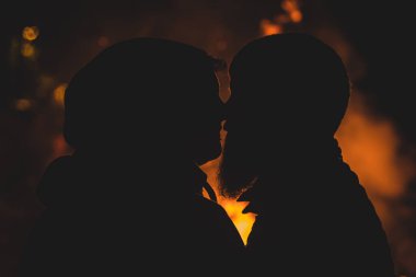 Ateşin önünde öpüşen ve burunlarıyla dokunan hippi bir çiftin romantik fotoğrafı. Kamp ateşi sırasında hipster aşıkların silueti. Romantik ortam, modern aşıklar..