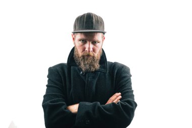 Beyaz stüdyo arka planında siyah kışlık ceket giyen sakallı, beysbol şapkalı sinirli görünüşlü bir hippi. Kızgın sakallı adam..