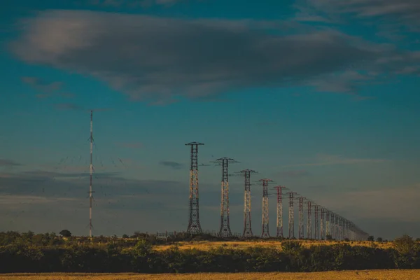 A row of pylons for power lines through a field in Ukraine. Next to it there is a radio tower antenna, on a hot summer evening.