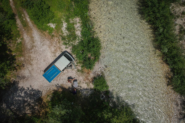 Wild camping next to a beautiful pristine crystal alpine river with an expeditional vehicle or old campervan. Drone aerial view of beautiful camping site.