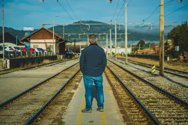 Man in jeans and jacket is waiting for a train in the early morning hours on a narrow train platform on a small train station.