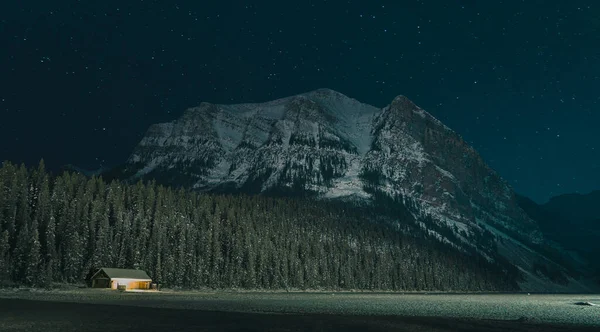 Small cabin on the banks of frozen Lake Louise during late winter night with stars rising above the mountain in the background.