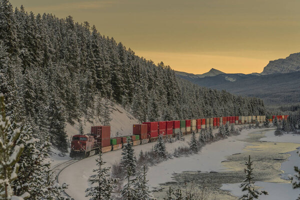 Freight train with containers after picturesque Morant's curve on a cold winter evening with sun just setting behind majestic mountains.