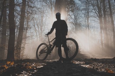 MTB or mountain biker posing in misty forest with sun behind him. Silhouette of a mountain biker in the foggy woods, beautiful sport silhouette clipart