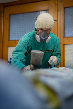 Yeşil tulumlu bir adam son bir kat boya için bir arabanın motor bölmesini hazırlıyor. Adam son boya kaplamasından önce araba panellerine gri sıvı macun veya sıva püskürtüyor..