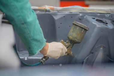 Yeşil tulumlu bir adam son bir kat boya için bir arabanın motor bölmesini hazırlıyor. Adam son boya kaplamasından önce araba panellerine gri sıvı macun veya sıva püskürtüyor..