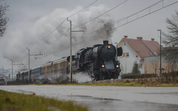 Old vintage steam train with black locomotive is running full power on a railroad track in the city during cold rainy season.