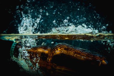 Kahverengi bira şişesi suya düşer ve büyük damlalar atar ve koyu bir arka plana püskürtür. Suyun içindeki taze ve sulu içecek konsepti.