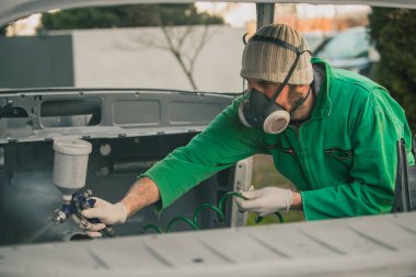Kafkasyalı adam, restorasyon projesi olarak arabanın üzerine sıkıştırılmış hava boyası tabancasıyla boya sıkıyor. Maske ve eldiven gibi koruyucu ekipmanlar giyen adam, profesyonel otomotiv ressamı..