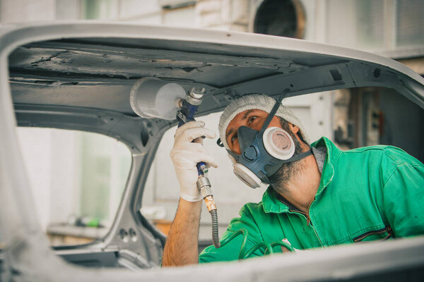 Caucasian man is spraying color with a compressed air paint gun on the vintage car as a restoration project. Man wearing protective equipment such as mask and gloves, professional automotive painter.
